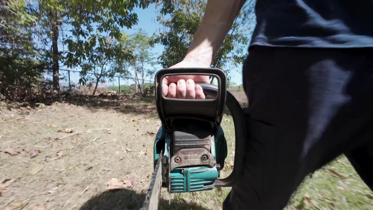 POV view of man carrying a chainsaw through an overgrown backyard, camera facing backwards against direction of travel. Property maintenance, vegetation clearing and land management concept.
