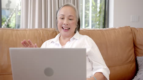 waving and smiling, elderly asian woman video chatting on laptop at home