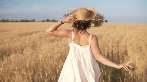 Seguimiento-De-Imágenes-De-Una-Hermosa-Chica-Con-Vestido-Blanco-De-Verano-Y-Sombrero-De-Paja-Corriendo-Libremente-Por-El-Campo-De-Trigo.-Vista-Trasera