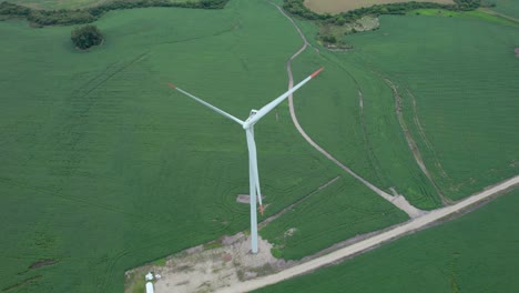 Toma-Aérea-De-Una-Turbina-Eólica-Solitaria-En-Un-Exuberante-Campo-Verde,-Camino-De-Acceso-De-Tierra-Visible,-Día-Nublado