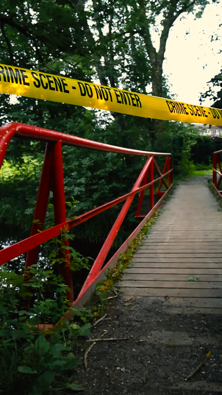 Video girato in verticale di un nastro giallo della polizia "Crime Scene Do Not Enter" teso davanti a un ponte rosso in campagna durante l'estate, con alberi e un fiume visibili sullo sfondo.