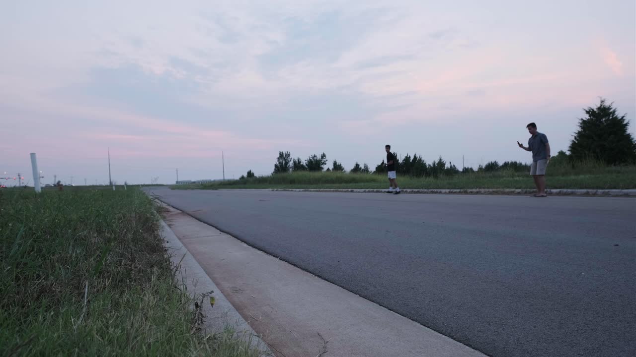 Wide static shot of friend recording a male skateboarder failing a skateboard trick on an empty road.