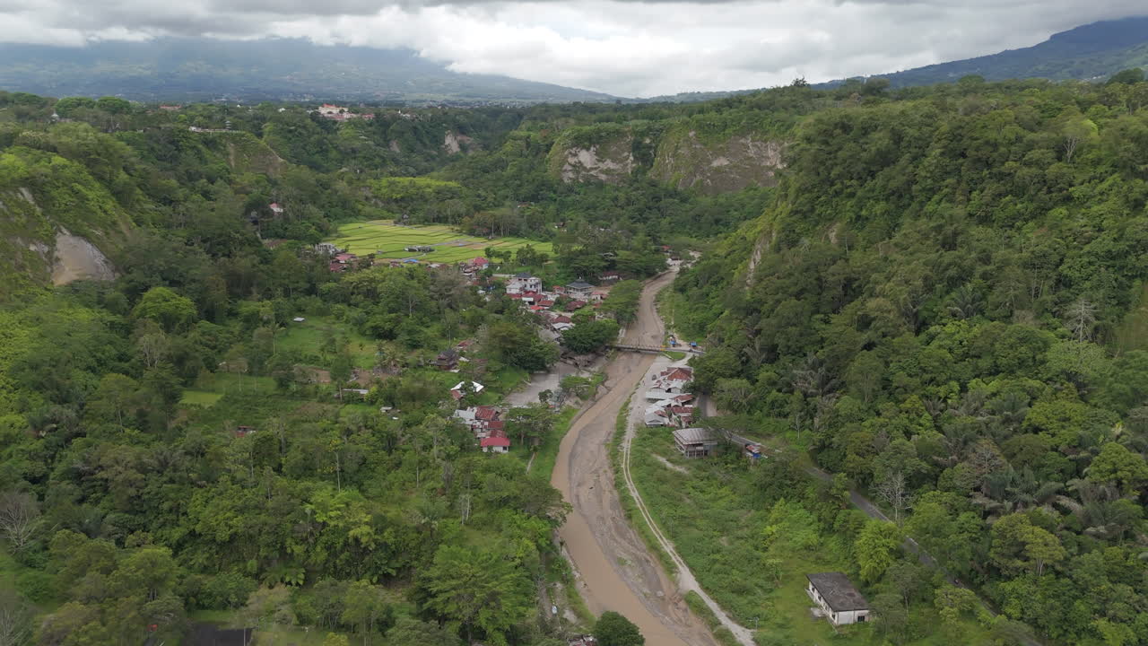 Aerial view descending into the deep Sianok Canyon (Ngarai Sianok), steep cliffs, a winding muddy river, lush tropical forest, and a small residential village below. West Sumatra, Indonesia