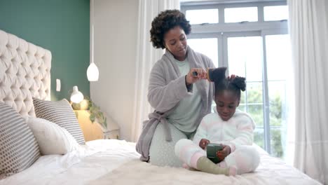 Glückliche-Afroamerikanische-Mutter-Kämmt-Die-Haare-Ihrer-Tochter-Mit-Dem-Smartphone-Im-Schlafzimmer,-Zeitlupe