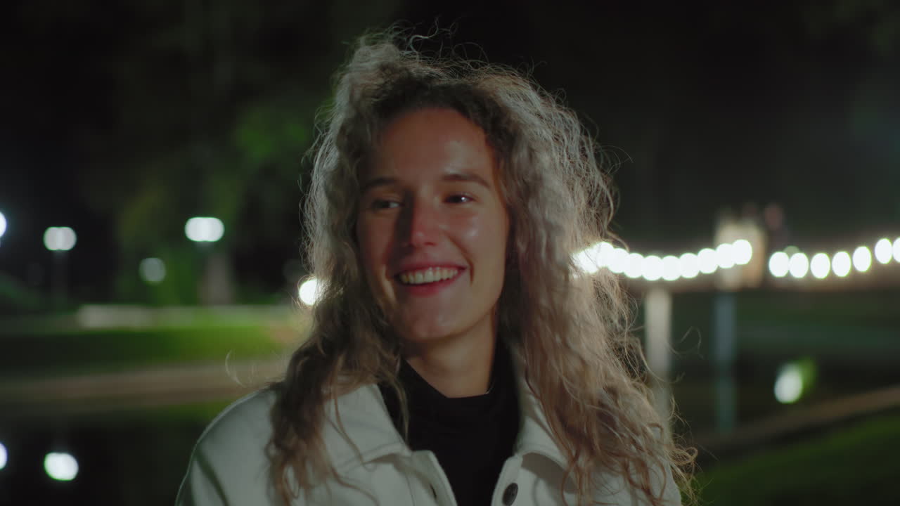Cheerful white woman with curly hair seated outdoors at night, hair swaying gently under glowing lamps, blur reflection of lights behind, radiating warm smile in relaxed atmosphere near calm water