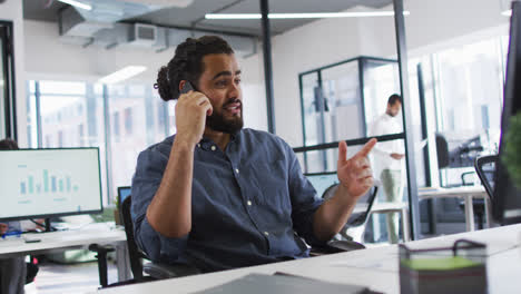 Mixed-race-businessman-sitting-at-desk-looking-at-computer-screen,-using-smart-phone-and-smiling