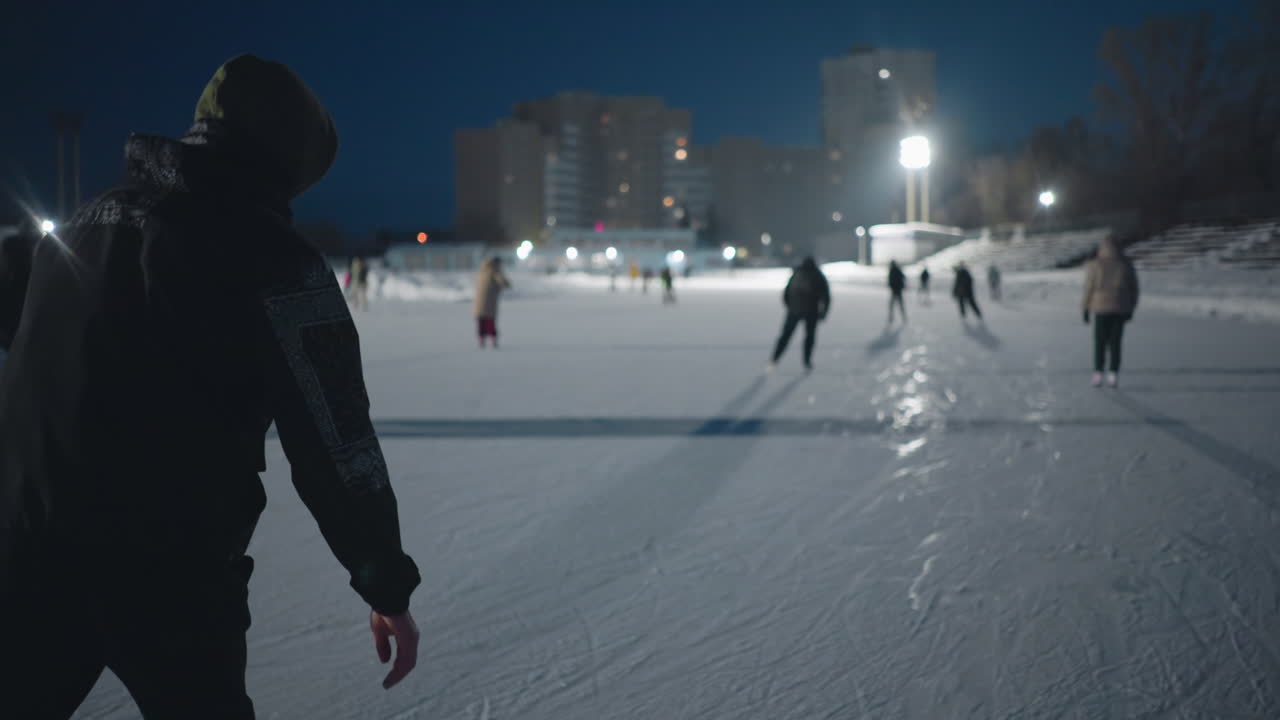 rear view student skating on outdoor ice rink under bright lights with several people skating around enjoying winter evening activity in urban setting blurred background features buildings