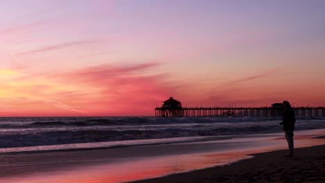 Un-Hombre-Disfruta-De-Sus-Vacaciones-En-La-Playa-Durante-Una-Hermosa-Puesta-De-Sol-Roja,-Púrpura,-Mandarina,-Rosa-Y-Azul-Con-El-Muelle-De-Huntington-Beach-Al-Fondo-En-Surf-City-USA-California