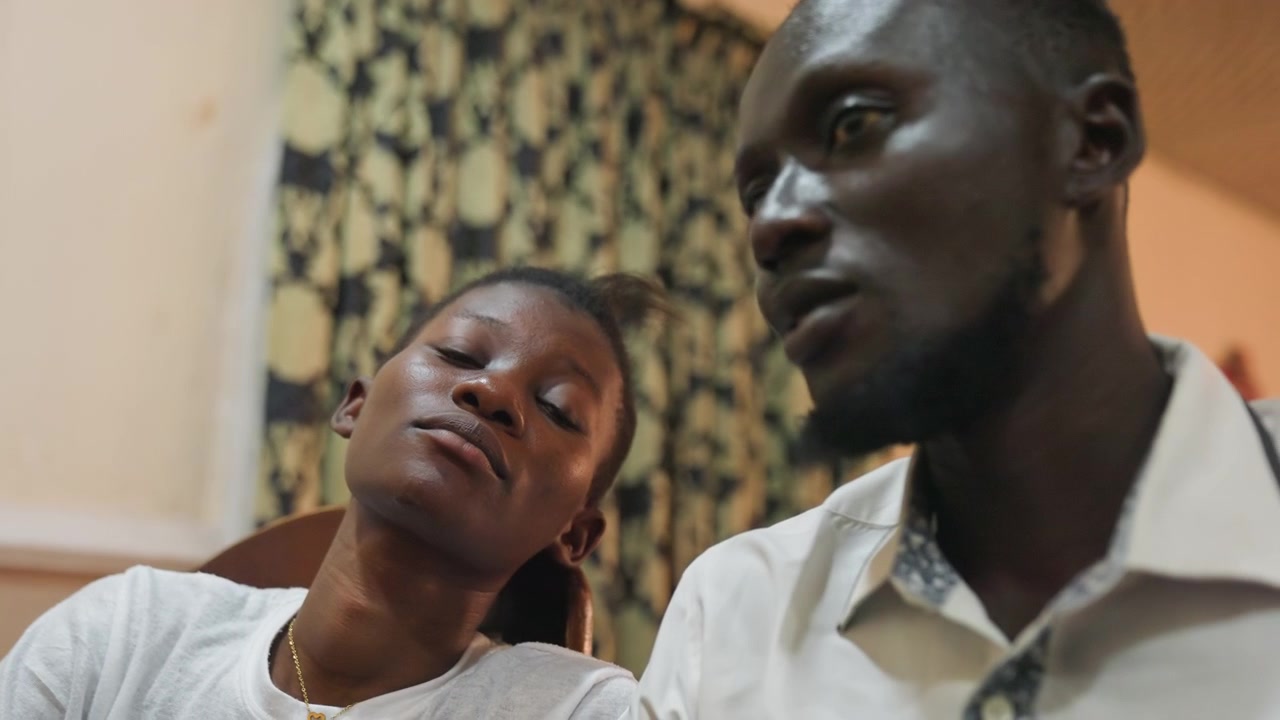 Couple noir curieux lors d'un rendez-vous intime, la femme observe l'homme avec un scepticisme amusé tandis qu'il répond avec un doux sourire, scène de salon intérieur avec un rideau à motifs, lumière chaude et tamisée, flirt candide, subtil