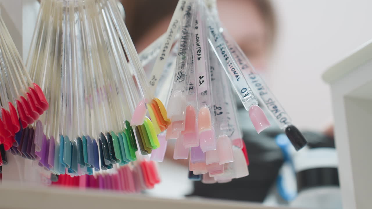 Close up view of colorful nail polish samples arranged on sticks with handwritten labels, hanging in foreground while focused nail technician works in blur background under soft lighting