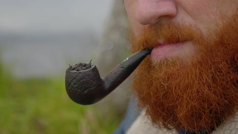 a close up shot of a ginger bearded man smoking a tobacco pipe and blowing out smoke