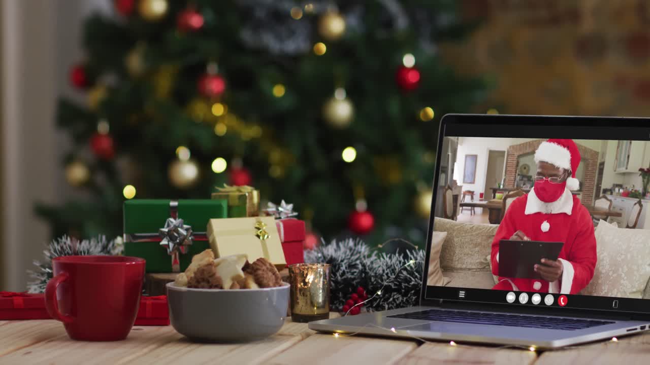 papá noel afroamericano con máscara facial en una videollamada en una computadora portátil, con decoraciones navideñas y árbol