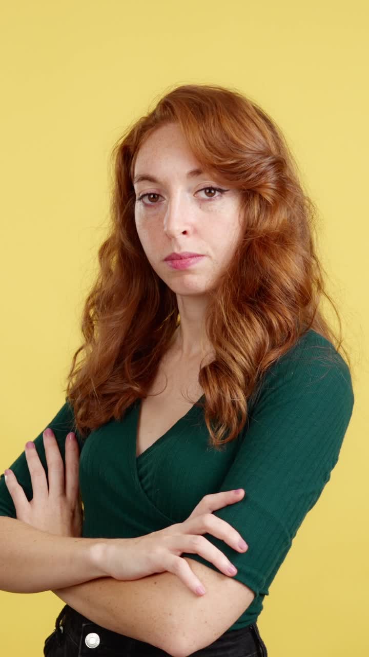 Serious woman with red hair and freckles crossing arms on yellow background