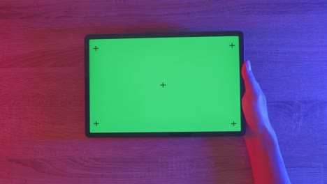 mano sosteniendo una tableta con una pantalla verde en una superficie de madera bajo luces de colores