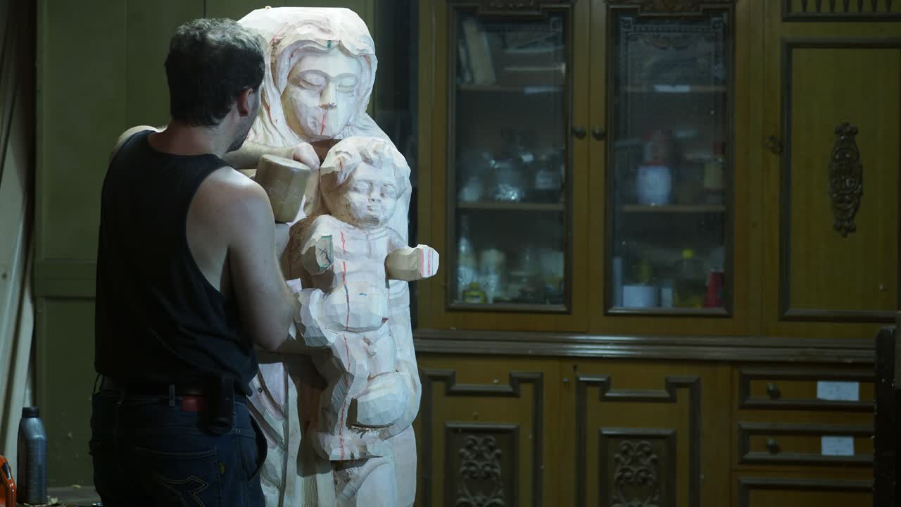 Craftsman hand-carves a wooden statue of the Virgin Mary holding baby Jesus, shaping details with chisels in a traditional workshop, showcasing patience, skill, and sacred artistry