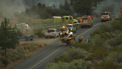 Aerial-fire-fighting,-helicopter-tackling-forest-fires-in-California-taking-pit-stop-on-rural-road,-firemen-around-aircraft-with-fire-trucks-in-background,-distant-view,-june-2020