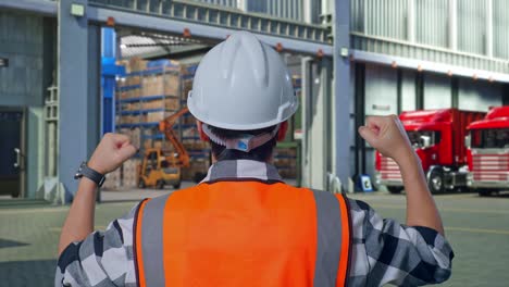 close up back view of a male engineer with safety helmet raising his hands celebrating while working , outside of logistics distributions warehouse
