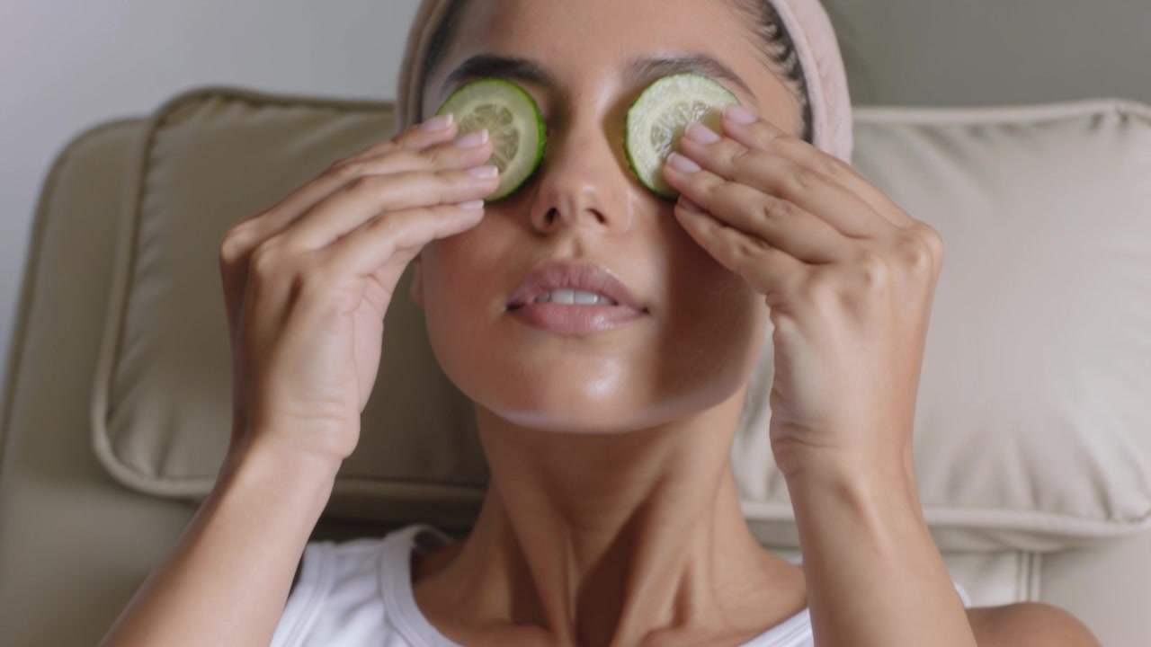Placing two green slices, woman covering closed eyes at home wearing pink headband for eye-care