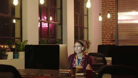 young caucasian business woman student working late using computer typing research project browsing online brainstorming deadline in trendy office at night wearing headphones