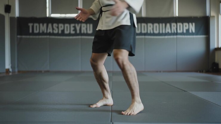 Responding to signal, practitioner in white gi and belt training footwork on gray mats at academy