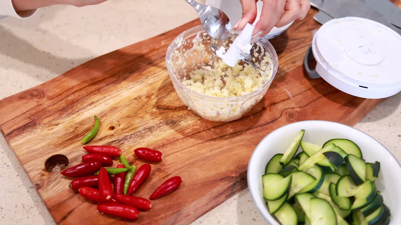 Hands using a manual chopper to mince garlic on a wooden board with fresh vegetables nearby