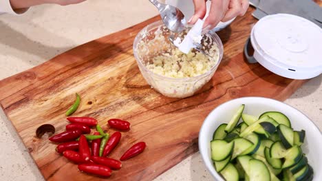 hands using a manual chopper to mince garlic on a wooden board with fresh vegetables nearby