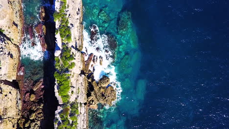 Drone-looking-straight-down-while-flying-over-long-narrow-rock-structure-and-sea-side-cliffs-surrounded-by-clear-blue-water