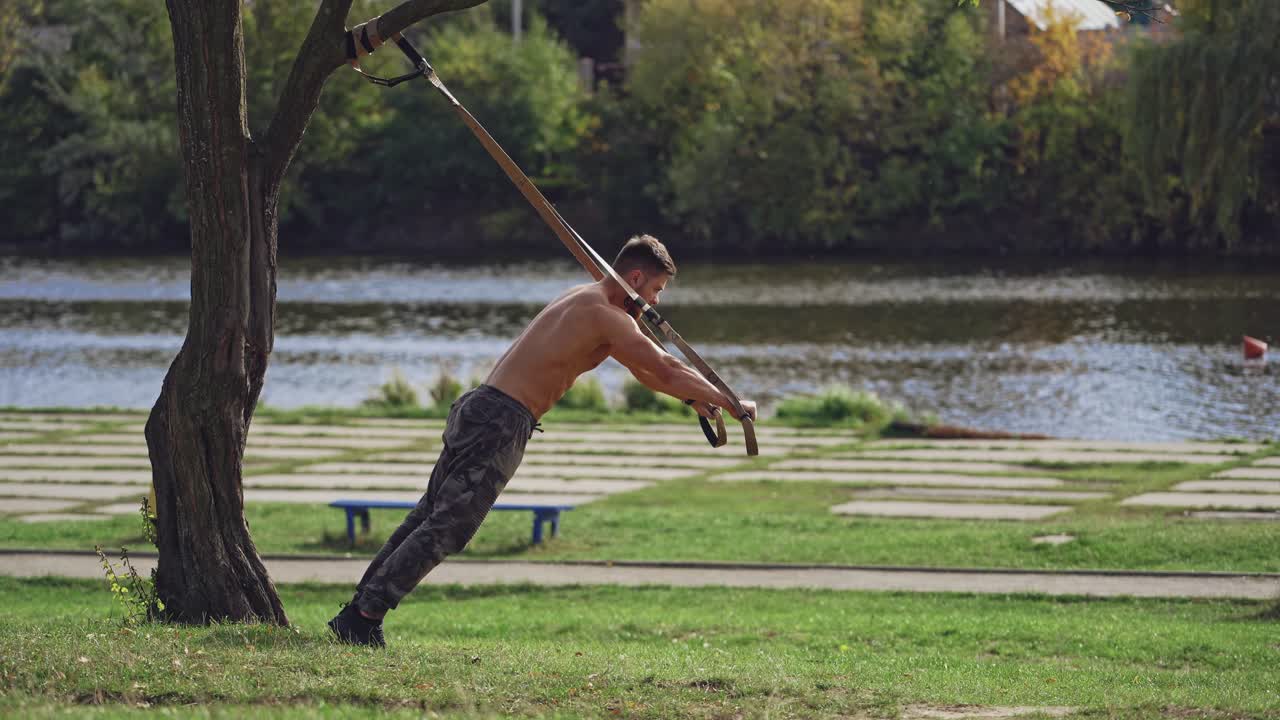 Young active man exercises outdoors with straps. Topless athlete doing workout on trx belts near the river in a summer day.