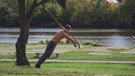 young active man exercises outdoors with straps. topless athlete doing workout on trx belts near the river in a summer day.