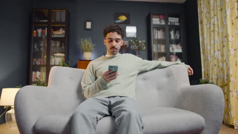 zoom out view of a young man using smartphone while sitting on a couch at home