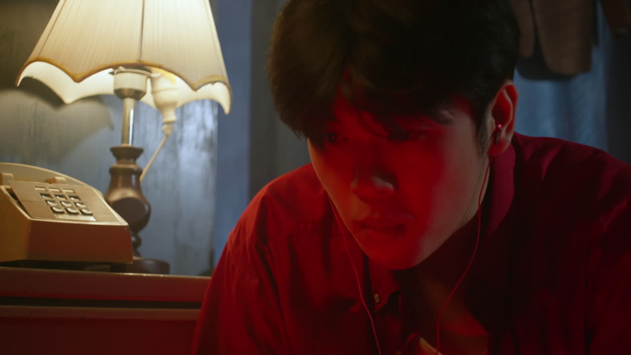 외로운 슬픈 아시아 남성이 호텔 방 침대에 혼자 앉아 헤드폰으로 음악을 듣고 있습니다.