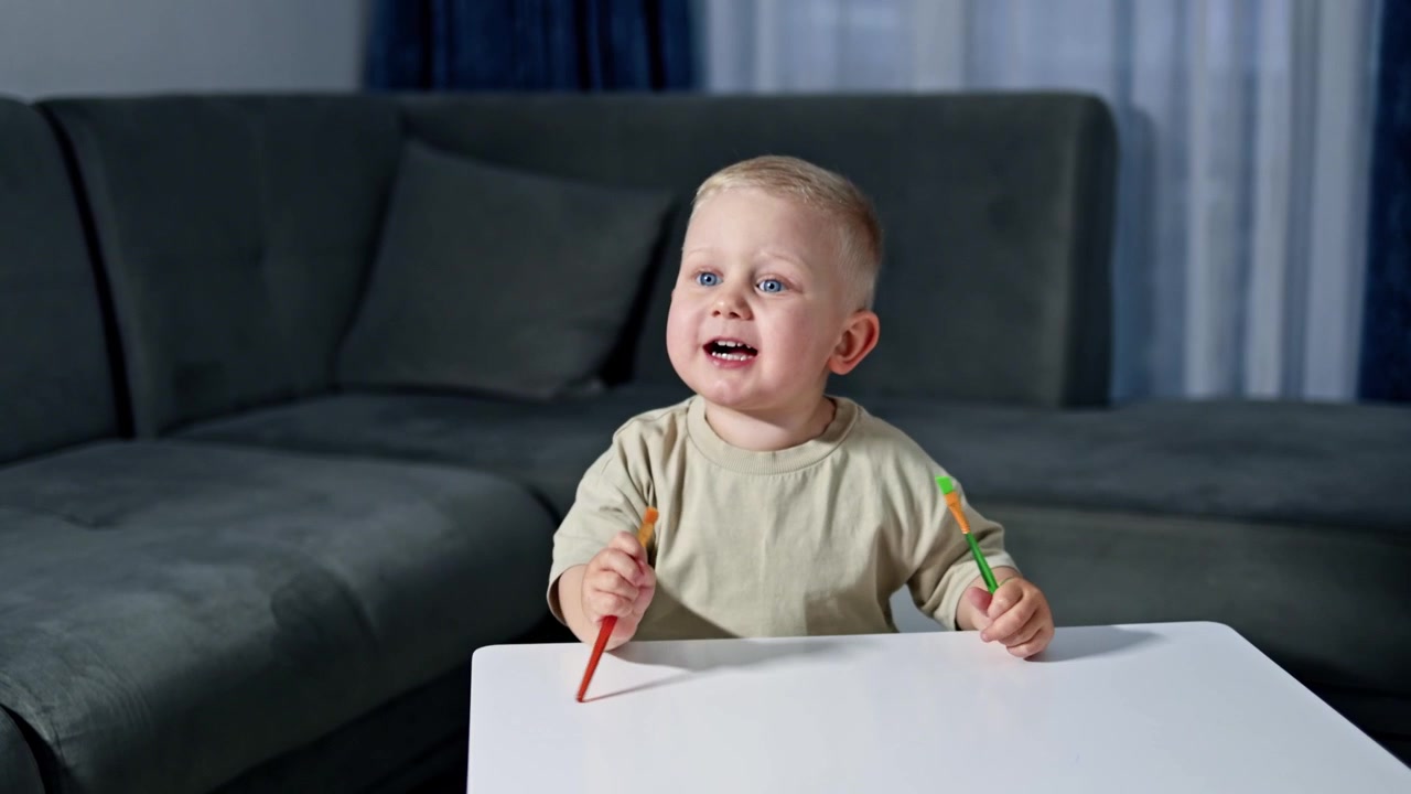 Süßes Kleinkind sitzt zu Hause und malt Bilder mit Farben. Kind bastelt und zeichnet auf Papier. Zweijähriger Junge hat Spaß.