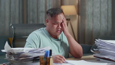 tired fat asian man putting hand on cheek while working hard with documents at the office
