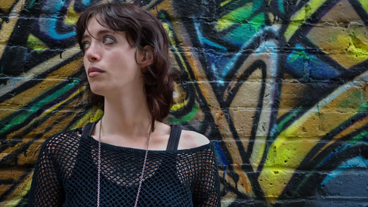 retrato de moda al aire libre de una mujer joven de estilo alternativo apoyándose en una pared cubierta de graffiti en la calle de la ciudad de londres, reino unido, en tiempo real.