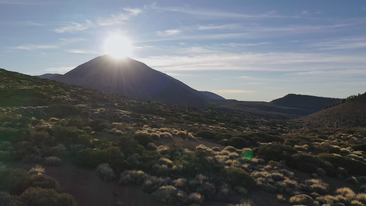 세 명의 등산객이 카나리아 제도의 피코 데 테이데 산 아래 언덕에 서서 바위와 덤불 사이 정상 뒤로 지는 해를 바라보고 있습니다.