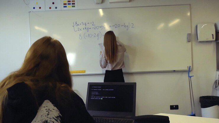 En ung kvindelig studerende ser sin lærer løse en matematisk ligning på en tavle i et klasseværelse, med en åben bærbar computer, der viser en programmeringsgrænseflade, på skrivebordet i forgrunden