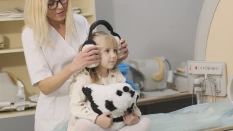 el médico radiólogo pone auriculares de aislamiento de ruido en el paciente niña para hacer un examen de resonancia magnética, la niña está sentada en la mesa automática