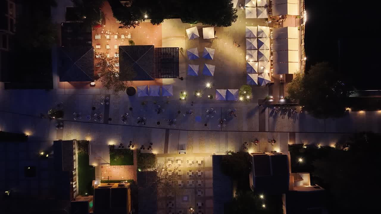 Top-down aerial (nadir) view of a resort plaza at night. People sitting at tables in an outdoor cafe or restaurant