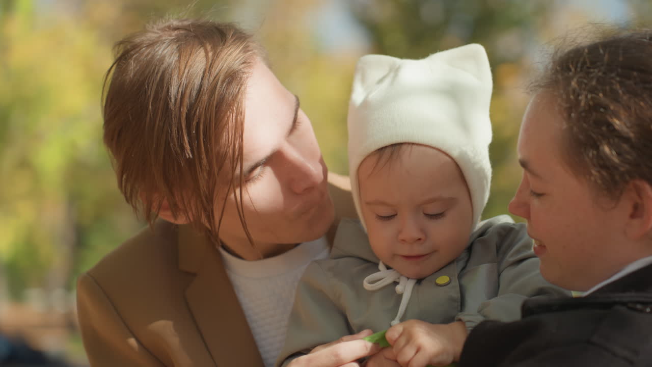 Baby wird von Eltern im Freien gehalten, junges Ehepaar führt neugieriges Kleinkind über einen Parkweg, Blatt- und Baumstammtexturen, weiches Sonnenlicht filtert durch, intime Elterngesten und schützender Blick.