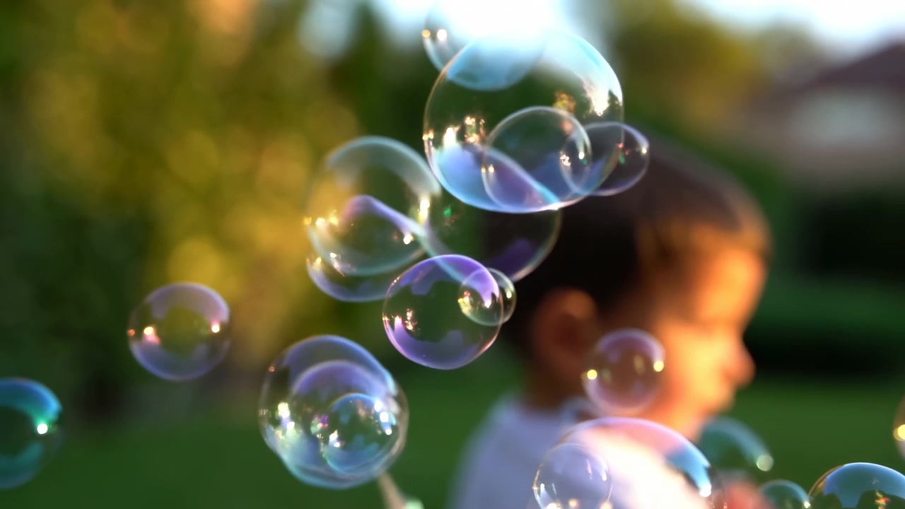 Translucent soap bubbles drift through foreground in layered clusters, catching bright rainbow highlights and glossy reflections while blurred child profile stays softly out of focus outdoors