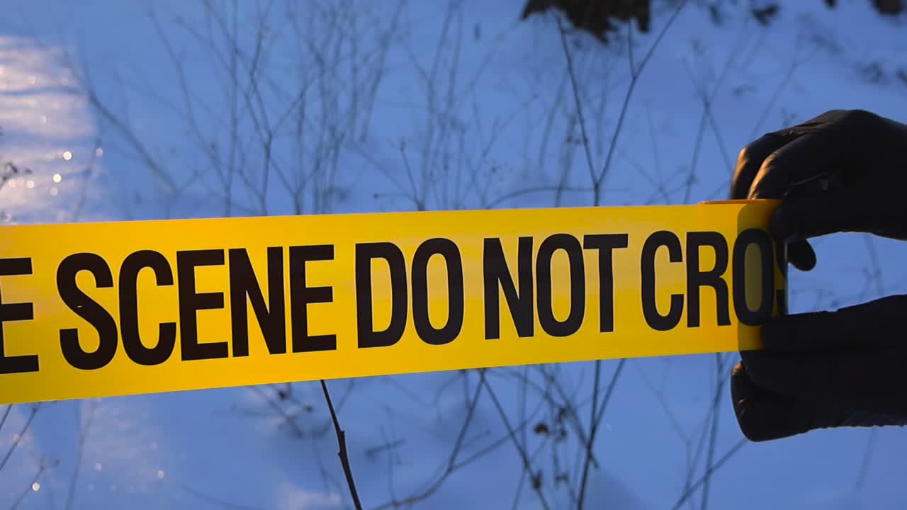 Close up of gloved investigator hands unrolling yellow crime scene tape in sunlit snowy forest with warm backlight and sparkling winter snow