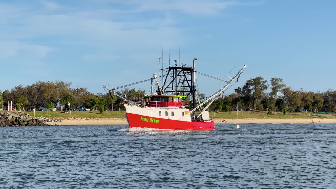 Red-hulled fishing trawler moves steadily along calm river under clear daylight, wide static shot