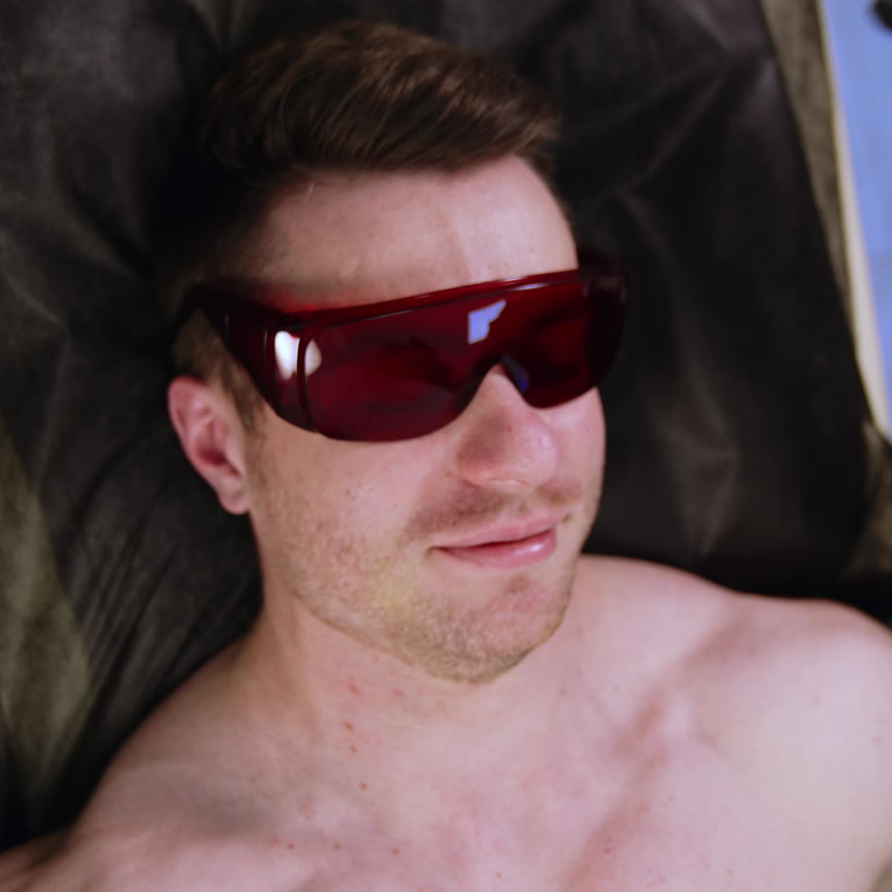 Mid-aged Caucasian man lies on the bed. Specialist in latex gloves puts protective eyeglasses on a male client