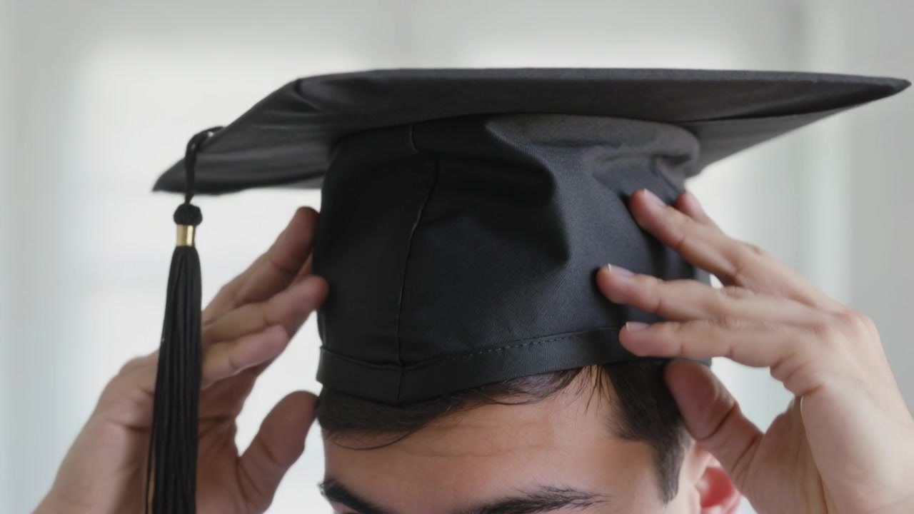 Uomo che tiene un tocco nero sopra la testa, sistemando il cappello in una stanza luminosa, mostrando la nappa e la camicia