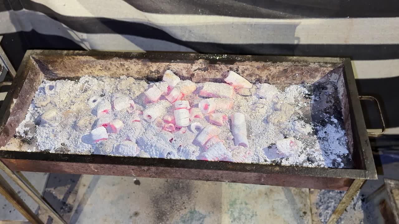 glowing charcoal embers covered in ash, heating up for grilling. Hot coals create a rustic outdoor cooking atmosphere with bright red heat and texture