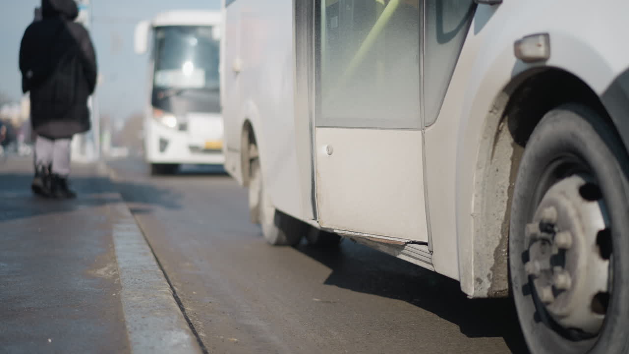 curb view shows bus stopped at stop with door open, passengers step off while new riders line up to board, wheel and pavement in focus, distant bus approaches, morning commute under cold light