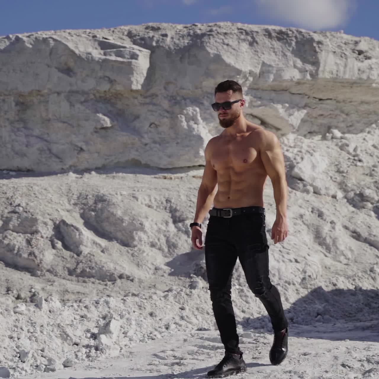 Handsome man near the black car. Shirtless guy in black sunglasses with sportive body walking in white mountains in a summer day. Slow motion.