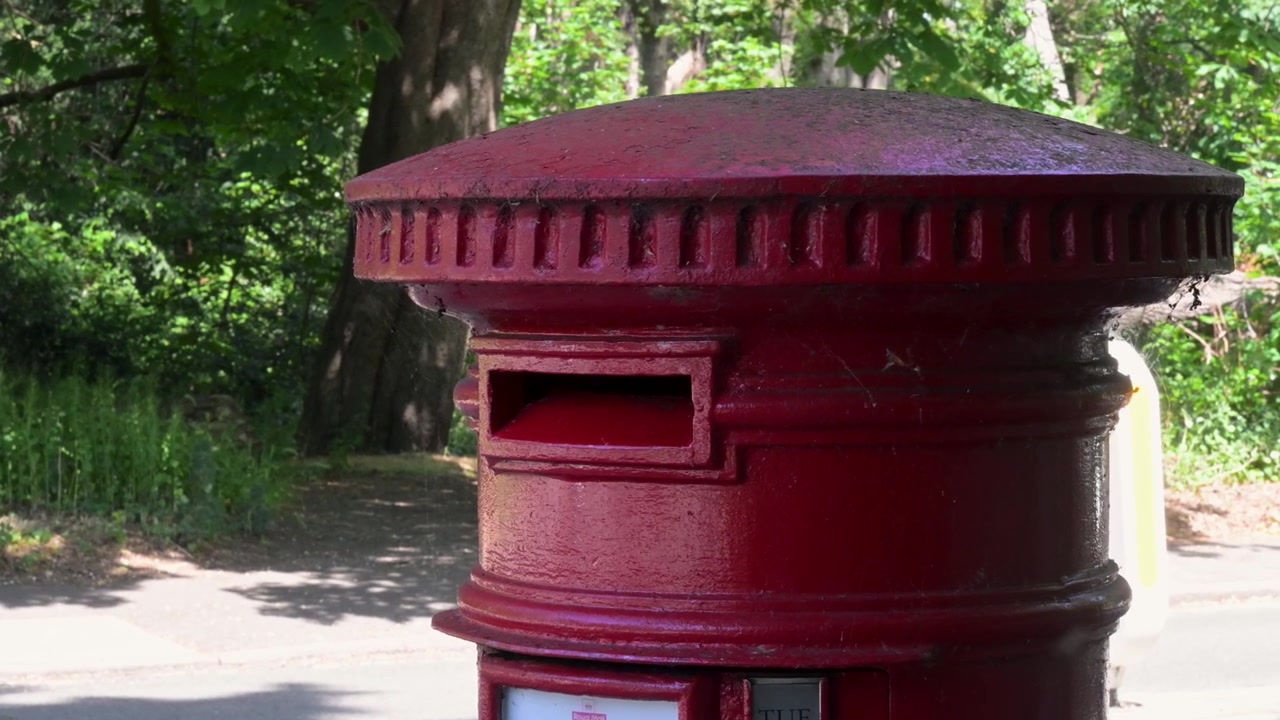 Un clásico buzón de correos rojo de pilar se alza contra un telón de fondo de árboles iluminados por el sol mientras los vehículos pasan. Captura la vida urbana y el encanto histórico. Ideal para temas urbanos e históricos.