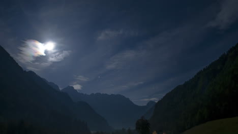 Timelapse-of-Alpine-valley-by-night-under-full-moon,-illuminated-farmhouse,-cottage,-full-moon,-clouds-and-stars-in-sky,-zoom-out-shot,-revealing