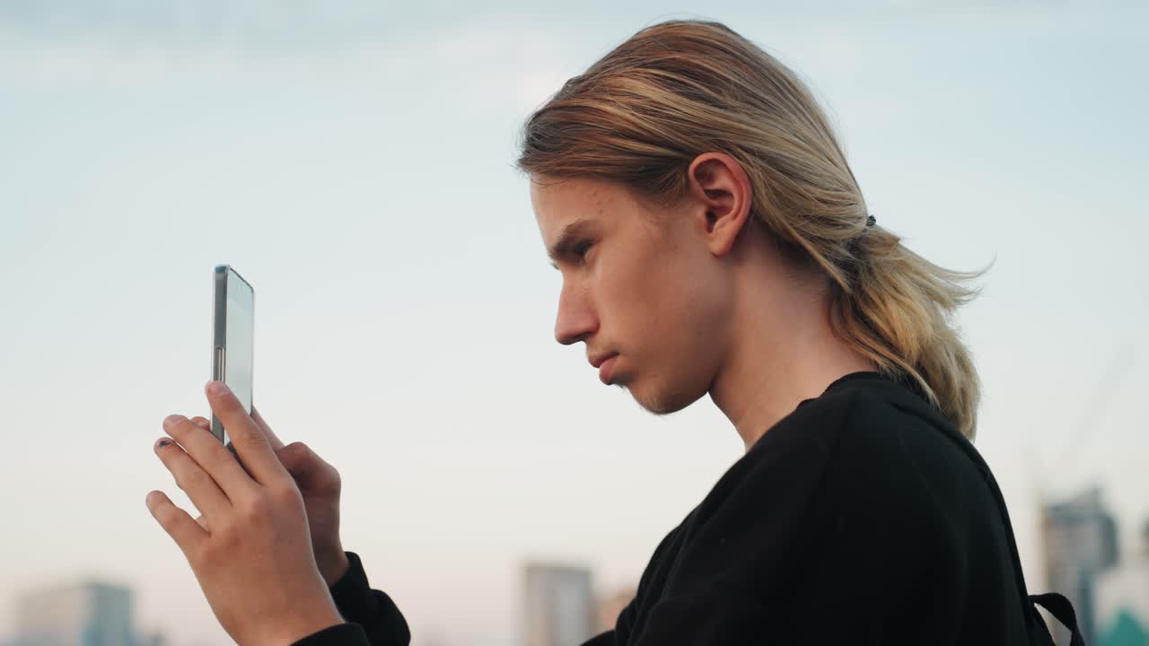Un adolescente esamina il dispositivo. Un giovane maschio ispeziona il suo dispositivo mobile mentre lo skyline della città si illumina al crepuscolo. Un giovane nella sua adolescenza esamina attentamente il suo smartphone con le luci urbane che incombono dietro di lui.